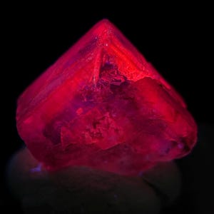 May include: A vibrant, faceted crystal with a deep red hue, illuminated against a black background. The gemstone's surface reflects light, showcasing its crystalline structure. The crystal sits on a light-colored base.