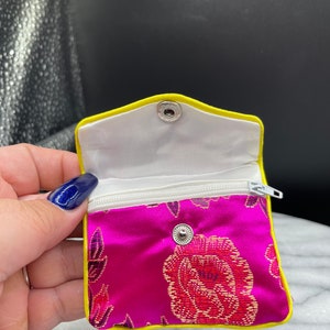 May include: A small pink satin pouch with a zipper closure and a yellow trim. The pouch has a floral pattern and a snap closure.