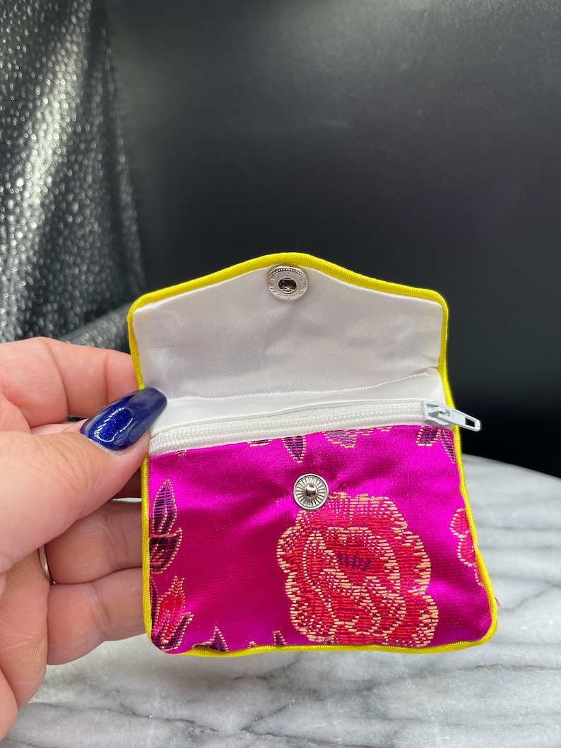May include: A small pink satin pouch with a zipper closure and a yellow trim. The pouch has a floral pattern and a snap closure.