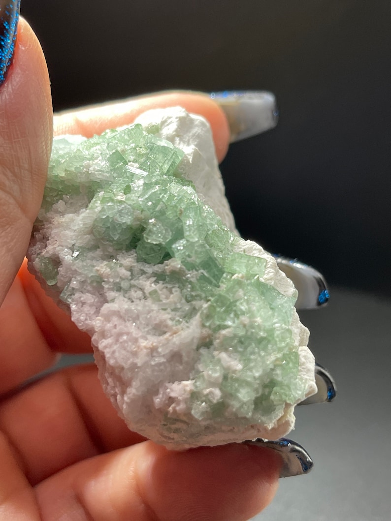 May include: A cluster of green crystals on a white matrix. The crystals are small and have a glassy lustre. The matrix is rough and has a white colour.