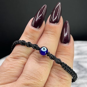 May include: A black woven bracelet featuring a blue and white evil eye charm. The bracelet is knotted and appears to be made of a durable cord. The charm is a deep blue color with a white and black eye design.
