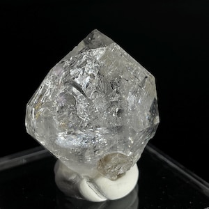 May include: A clear, faceted, quartz crystal with a double termination. The crystal is sitting on a white pedestal.