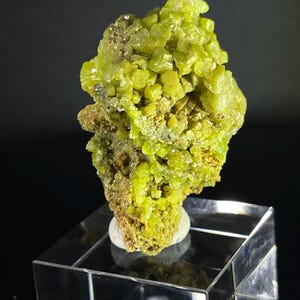 May include: A vibrant, lime-green mineral specimen with a crystalline structure. The mineral sits atop a clear, geometric display stand, showcasing its natural form. The background is a dark, solid color, highlighting the bright green hue of the mineral.
