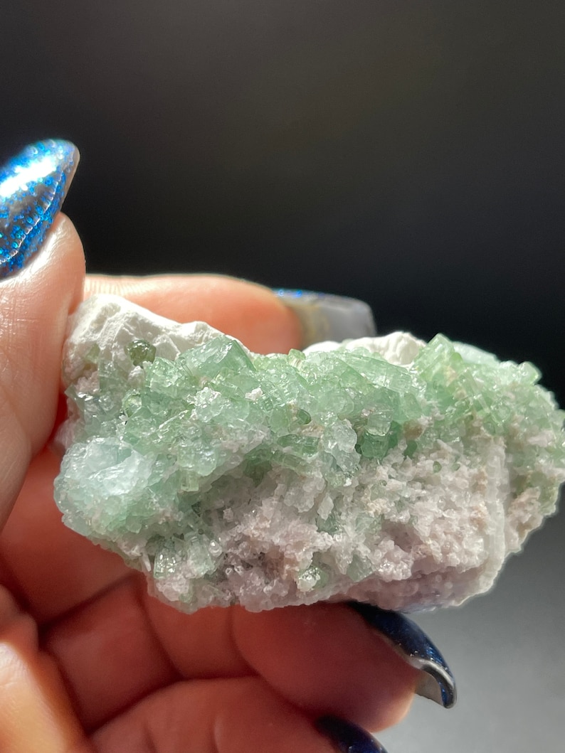 May include: A cluster of green crystals on a white matrix. The crystals are small and have a glassy lustre. The matrix is rough and has a white colour.