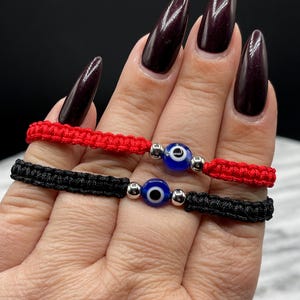 May include: Two woven bracelets, one red and one black, each featuring a blue evil eye bead with silver accents. The bracelets are displayed on a hand, showcasing their intricate design and vibrant colors. A close-up shot highlights the details.