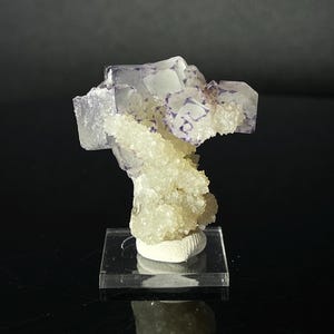 Peut inclure: Un groupe de cristaux de fluorine violets avec des cristaux de calcite blancs sur un support d'exposition transparent.