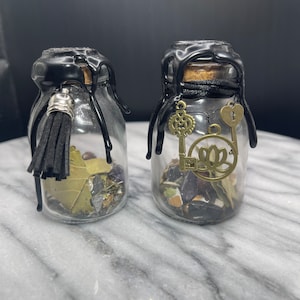 May include: Two small glass bottles with black wax seals and cork stoppers. Each bottle contains various herbs, crystals, and charms. One bottle has a black tassel and the other has a key and a lotus charm.