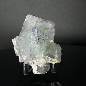 Peut inclure: Un amas de cristaux de fluorite translucides aux teintes vert pâle et violettes. Le spécimen minéral est monté sur un présentoir noir, mettant en valeur sa structure cristalline naturelle. Le fond est gris foncé.