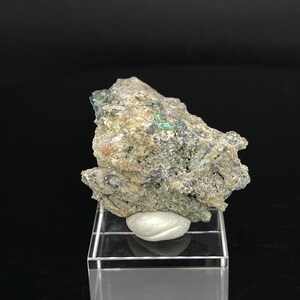 May include: A rough, gray rock with green and blue mineral inclusions. The rock is displayed on a clear acrylic stand.
