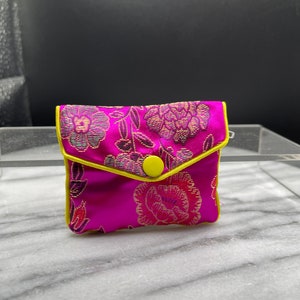 May include: A small pink pouch with a floral pattern and a yellow button. The pouch is lined with a yellow trim.