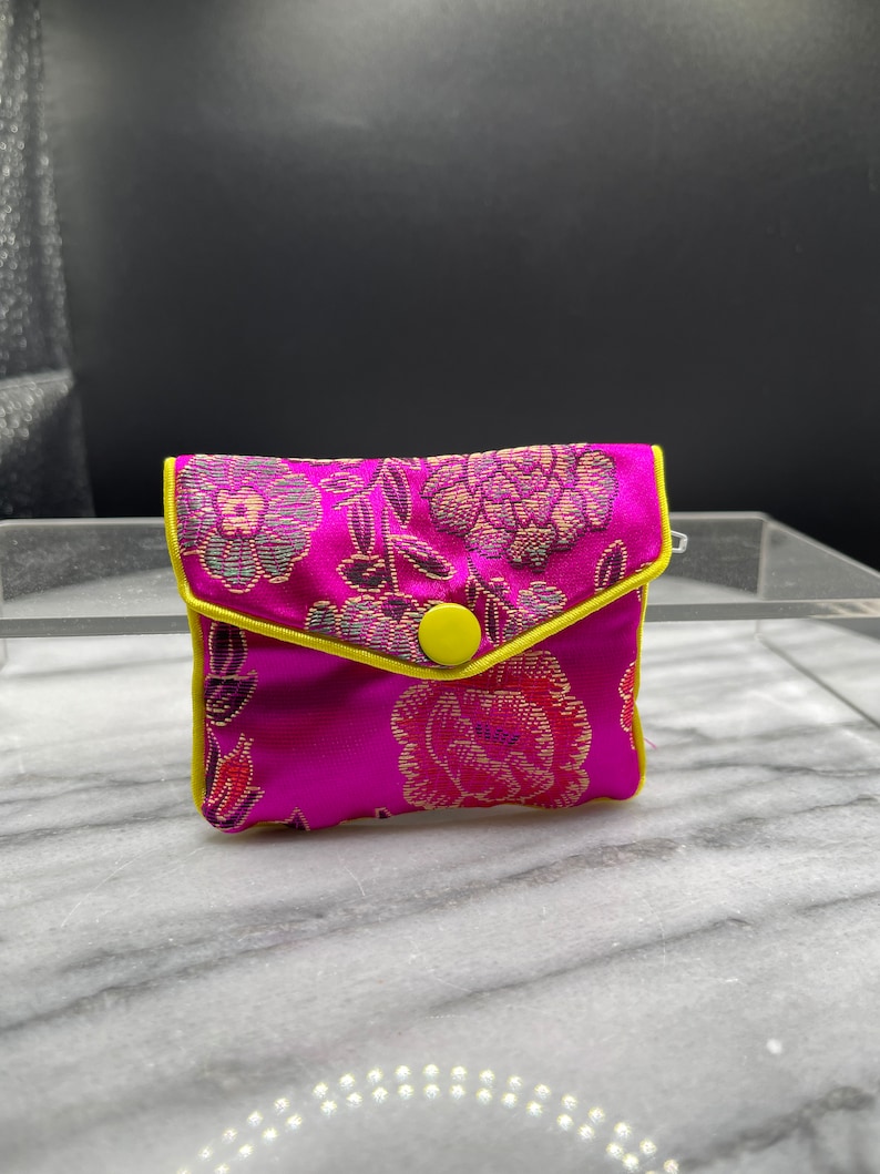May include: A small pink pouch with a floral pattern and a yellow button. The pouch is lined with a yellow trim.