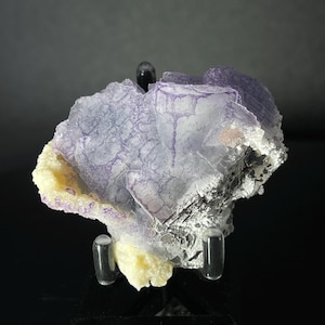 Peut inclure: Un cristal de fluorine violet et blanc avec une base de calcite jaune. Le cristal est sur un support noir.