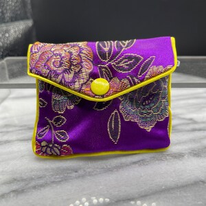 May include: A small purple pouch with a yellow button closure and floral pattern. The pouch is made of a soft, silky fabric.