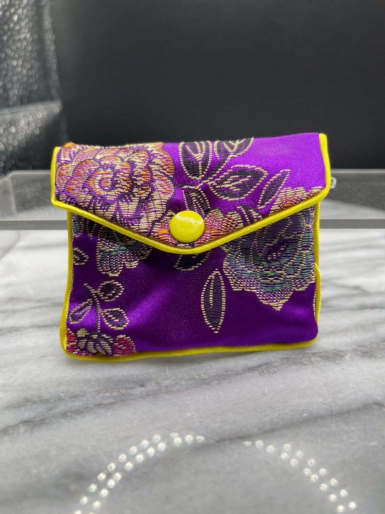 May include: A small purple pouch with a yellow button closure and floral pattern. The pouch is made of a soft, silky fabric.