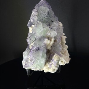 Peut inclure: Un groupe de cristaux de fluorine verts et violets sur une matrice blanche. Les cristaux sont disposés en forme de pyramide et sont exposés sur un support noir.