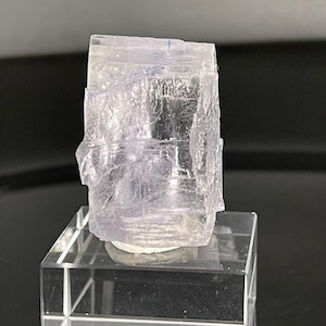 Puede incluir: Un cristal translúcido de color lavanda pálido con forma cúbica, exhibido en un soporte de vidrio transparente.