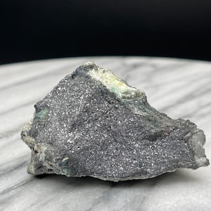 Può includere: Una roccia grigio scuro con un'inclusione minerale verde e bianca. La roccia è appoggiata su una superficie di marmo bianco.