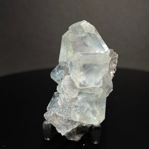 May include: A cluster of clear, translucent fluorite crystals with a slight blue hue. The crystals are arranged in a pyramid shape and have a smooth, glassy surface. The fluorite is displayed on a black stand.