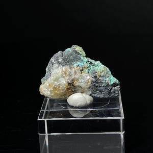 May include: A mineral specimen featuring a dark, textured rock with patches of green and blue hues. A small, oval, light-colored stone sits on a clear acrylic display stand. The background is black.