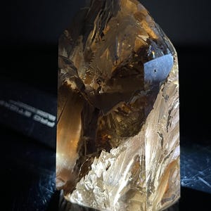 May include: A faceted, amber-colored crystal point with a reflective surface. The crystal has a rough, textured appearance with multiple facets and a pointed top. The lighting creates highlights and shadows, enhancing its crystalline structure.