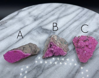 May include: Three pink and grey mineral specimens labelled A, B, and C on a white marble surface.