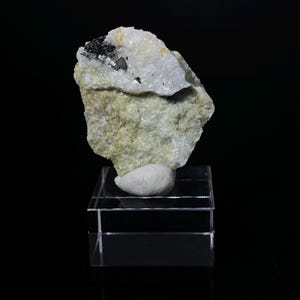 May include: A small, white and black mineral specimen on a clear acrylic display stand. The mineral has a rough, textured surface and is composed of several crystals.