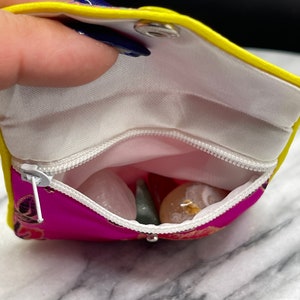 May include: A small pink pouch with a yellow zipper and a yellow flap. The pouch is open and contains several small stones, including a green stone, a white stone, and a pink stone.