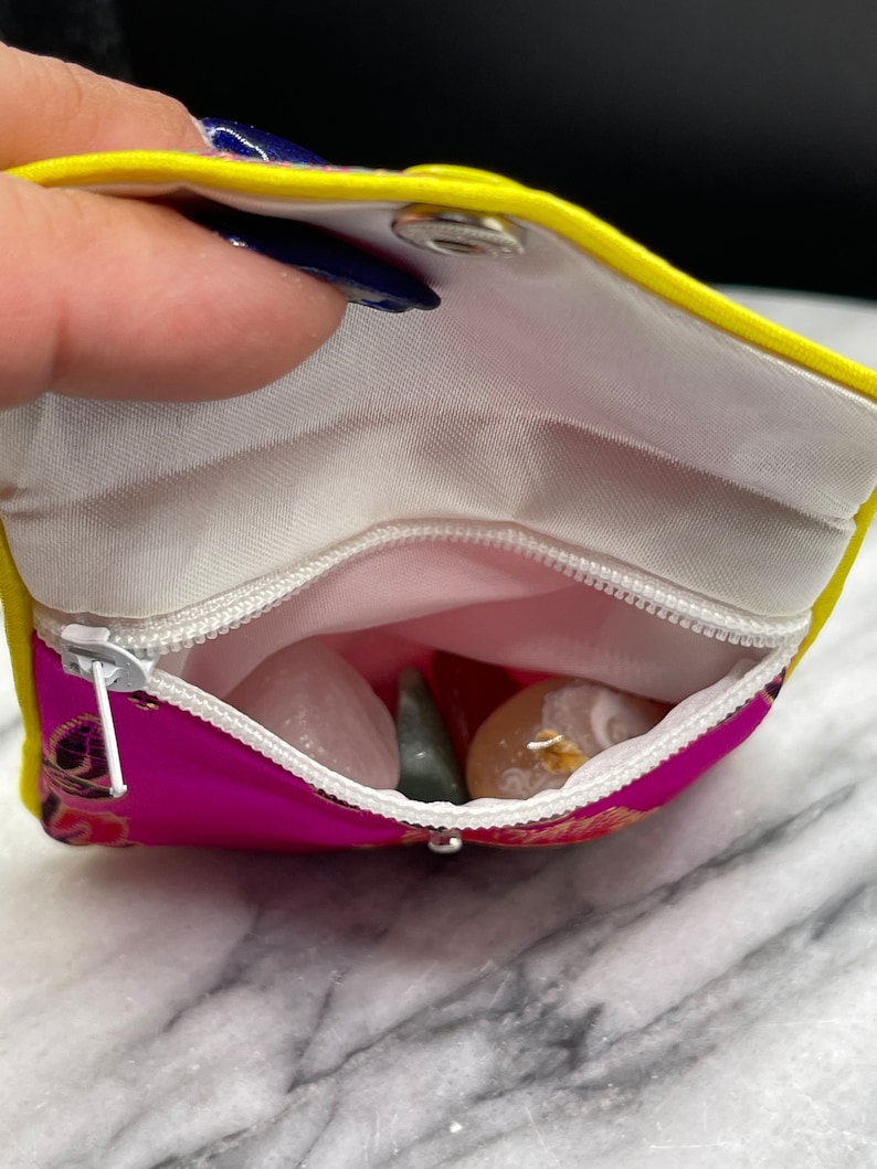 May include: A small pink pouch with a yellow zipper and a yellow flap. The pouch is open and contains several small stones, including a green stone, a white stone, and a pink stone.