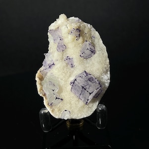 Peut inclure: Un groupe de cristaux de fluorine violets sur une matrice de calcite blanche. Les cristaux sont nets et bien définis, et la matrice est lisse et polie. L'échantillon est présenté sur un support noir.