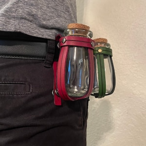 May include: Two glass bottles with cork stoppers, held in a leather carrier. One bottle has a burgundy leather strap, the other has a green strap. The carrier is attached to a black belt.