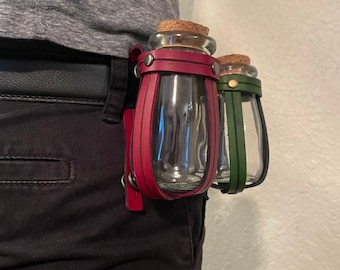 Puede incluir: Dos botellas de vidrio con tapones de corcho, sujetas en un soporte de cuero. Una botella tiene una correa de cuero burdeos, la otra una correa verde. El soporte está sujeto a un cinturón negro.
