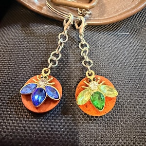 May include: Two keychains with orange circular pendants. Each pendant features a jeweled bee design. One bee has blue wings, the other has green wings. The keychains have silver chains and clasps.