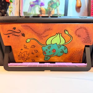 Può includere: Console Nintendo Switch nera con retro in pelle arancione personalizzato con un design Bulbasaur. Il Bulbasaur è verde e blu, con foglie e accenti a tema autunnale. La console è posizionata su un supporto nero.