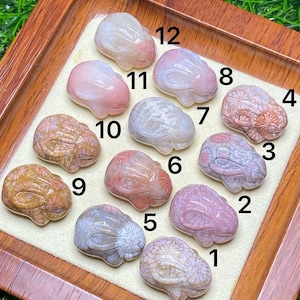 May include: A collection of twelve carved gemstone cabochons in various colors, including pink, white, and brown. Each stone has a unique pattern and is displayed on a wooden tray with a cream-colored insert. The gemstones are numbered 1 through 12.