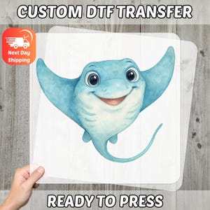 May include: A ready-to-press DTF transfer with a smiling cartoon stingray in blue and white. The image has the text "CUSTOM DTF TRANSFER" at the top and "READY TO PRESS" at the bottom. The transfer is displayed against a wooden background.