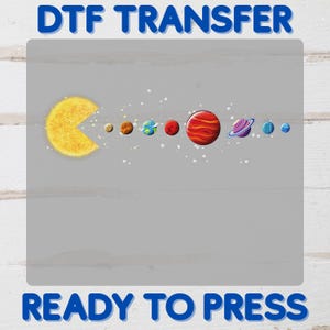 May include: A DTF transfer featuring a yellow sun-shaped Pac-Man eating its way through a colorful solar system. The planets include brown, blue, red, and striped designs. The text "DTF TRANSFER" and "READY TO PRESS" are in blue.
