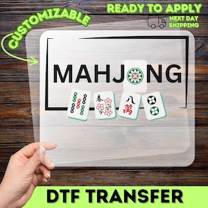 May include: A clear DTF transfer sheet featuring the word "MAHJONG" in bold black letters, with several Mahjong tiles. The transfer is labeled "CUSTOMIZABLE" and "READY TO APPLY". The background is a wooden surface.