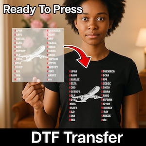 May include: Black t-shirt with a white airplane graphic and the phonetic alphabet. The words "Ready To Press" and "DTF Transfer" are displayed. The image is of a woman holding a clear sheet with the same design.