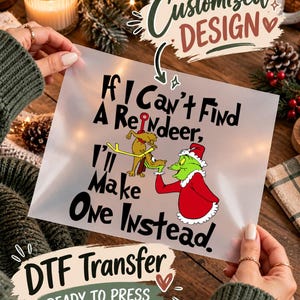 May include: A clear DTF transfer sheet featuring a graphic of a green character in a red Santa suit holding a small brown reindeer. The text reads, If I Can't Find A Reindeer, I'll Make One Instead. The design is printed in black, red, and green ink.