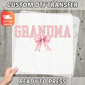 May include: A DTF transfer sheet featuring the word "GRANDMA" in pink glitter lettering and a pink bow. The text "CUSTOM DTF TRANSFER" and "READY TO PRESS" are at the top and bottom. The image is held by a hand.
