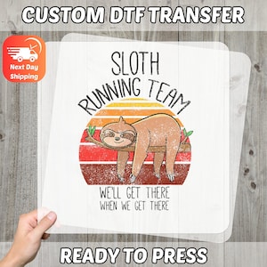 May include: A white DTF transfer featuring a sloth illustration on a branch, set against a sunset design. Text reads "SLOTH RUNNING TEAM" and "WE'LL GET THERE WHEN WE GET THERE". The transfer is labeled "READY TO PRESS".
