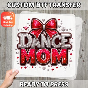 May include: A ready-to-press DTF transfer featuring the words "DANCE MOM" in a glittery red and silver design, with a red bow at the top. The transfer is on a clear sheet, held by a hand. The text "CUSTOM DTF TRANSFER" and "READY TO PRESS" are also visible.