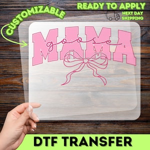 May include: A pink DTF transfer with the text "MAMA goose" in a playful font, featuring a bow design. The transfer is on a clear sheet, with the words "Customizable" and "Ready to Apply" visible. The background is a wooden surface.