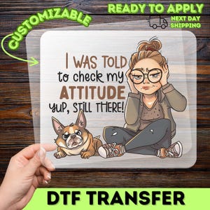 May include: DTF transfer with a cartoon of a woman and a dog, with the text "I WAS TOLD to check my ATTITUDE Yup, Still There!". The design is surrounded by the words "Customizable" and "Ready to Apply".