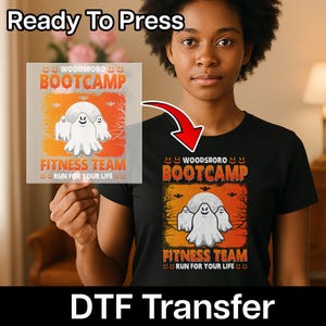May include: A black t-shirt featuring a graphic print with three white ghosts, a full moon, and bare trees against an orange background. The text reads WOODSBORO BOOTCAMP FITNESS TEAM RUN FOR YOUR LIFE, surrounded by small orange jack-o'-lantern faces.