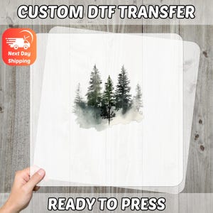 May include: A clear, rectangular DTF transfer sheet featuring a watercolor illustration of a forest with green and gray trees. The text "CUSTOM DTF TRANSFER" and "READY TO PRESS" are printed on the image.