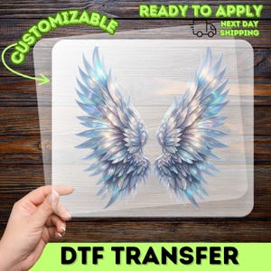 May include: A DTF transfer featuring a pair of detailed, iridescent angel wings in shades of silver and blue. The wings are displayed on a clear, rectangular sheet, with the words "CUSTOMIZABLE" and "READY TO APPLY" in green and white.