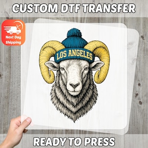 May include: A DTF transfer featuring a detailed illustration of a ram wearing a blue beanie with "LOS ANGELES" text. The ram has large yellow horns and a fluffy gray and white face. The words "CUSTOM DTF TRANSFER" and "READY TO PRESS" are also visible.