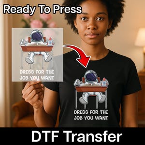 May include: Black t-shirt featuring an astronaut graphic at a school desk, a rocket, and the text "DRESS FOR THE JOB YOU WANT." The image also shows a DTF transfer and the text "Ready To Press."