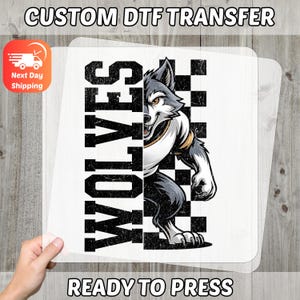 May include: A DTF transfer featuring a stylized wolf mascot with the word "WOLVES" in bold, black letters. The wolf is set against a black and white checkered background. The words "CUSTOM DTF TRANSFER" and "READY TO PRESS" are also visible.